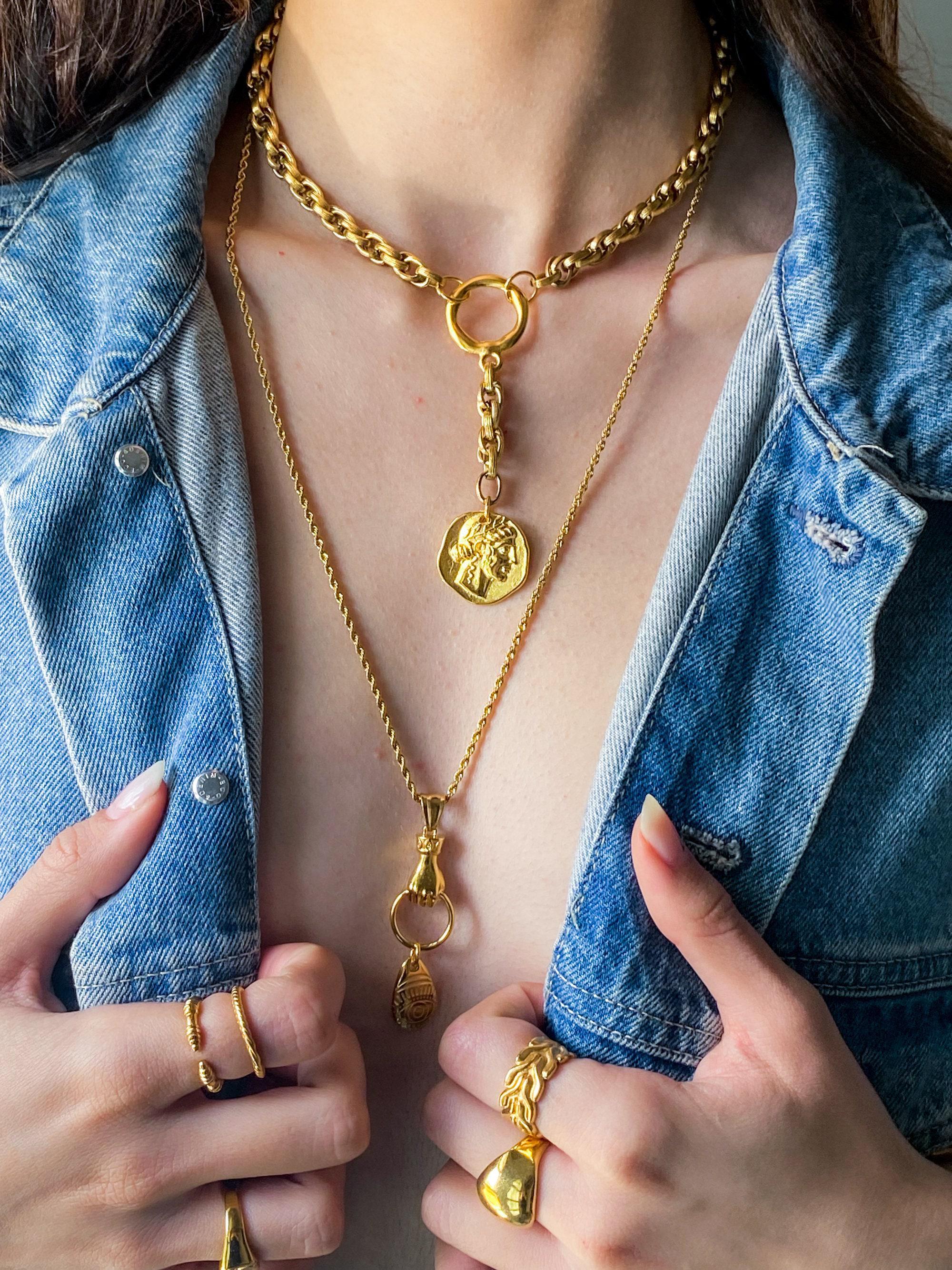 May include: Gold-colored necklaces and rings. The necklaces include a chain choker with a circular pendant, a coin pendant, and a hand pendant. The rings include a double-band ring and a leaf-shaped ring. The jewelry is displayed against a denim jacket.