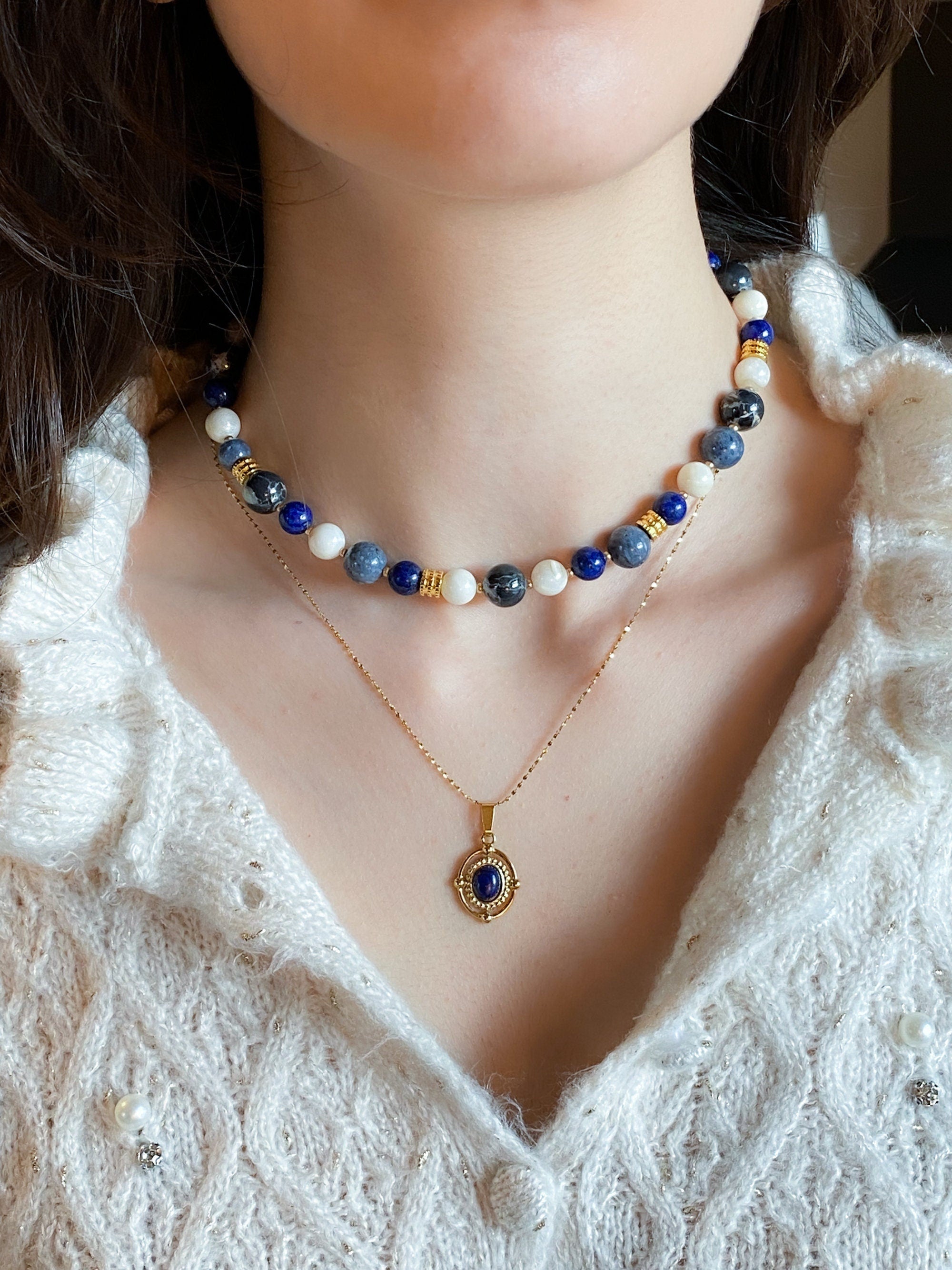 May include: A gold chain necklace with a blue gemstone pendant. The pendant is set in a gold frame with a detailed design. The necklace is layered over a beaded choker necklace with white, blue, and gray beads.