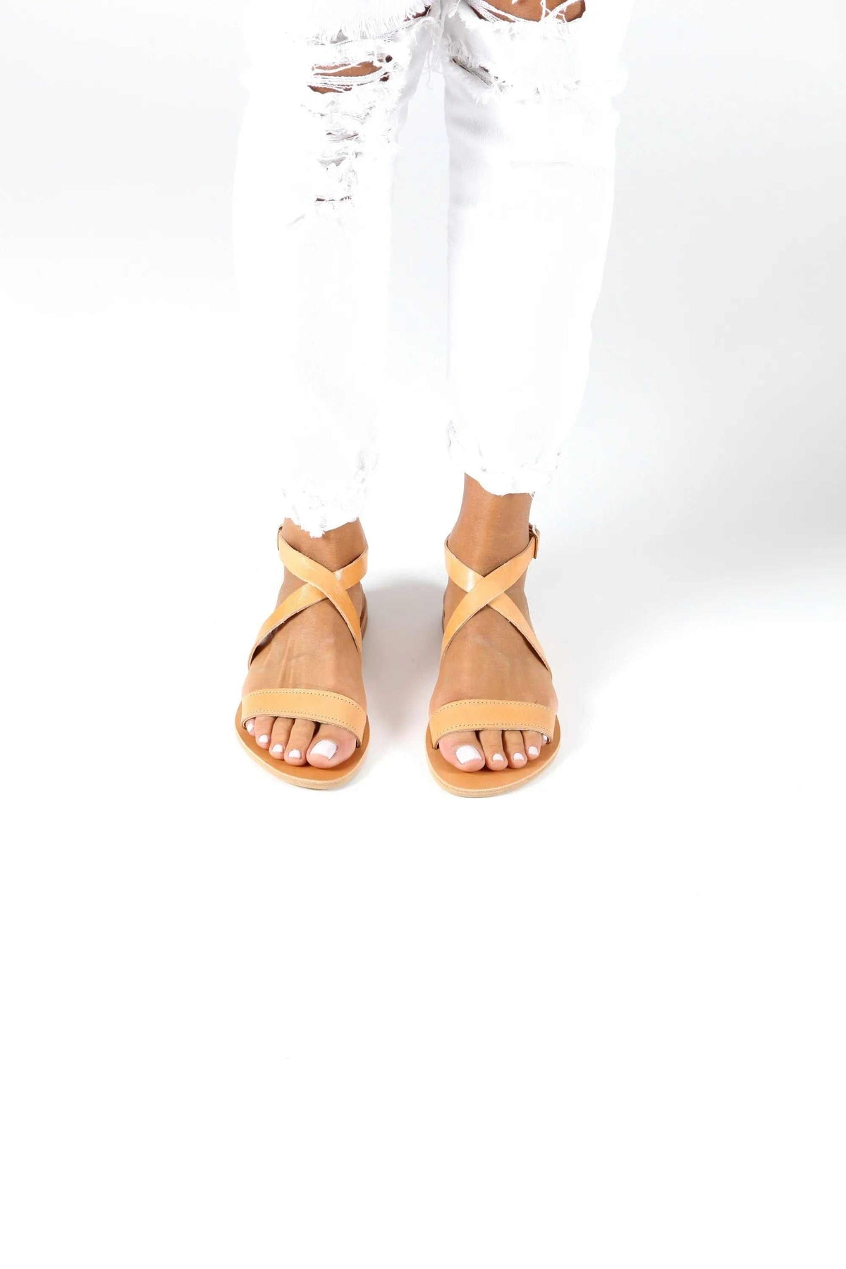 MYRTO Handmade Greek Sandals in tan leather with flat soles and crossed ankle straps, worn with white trousers
