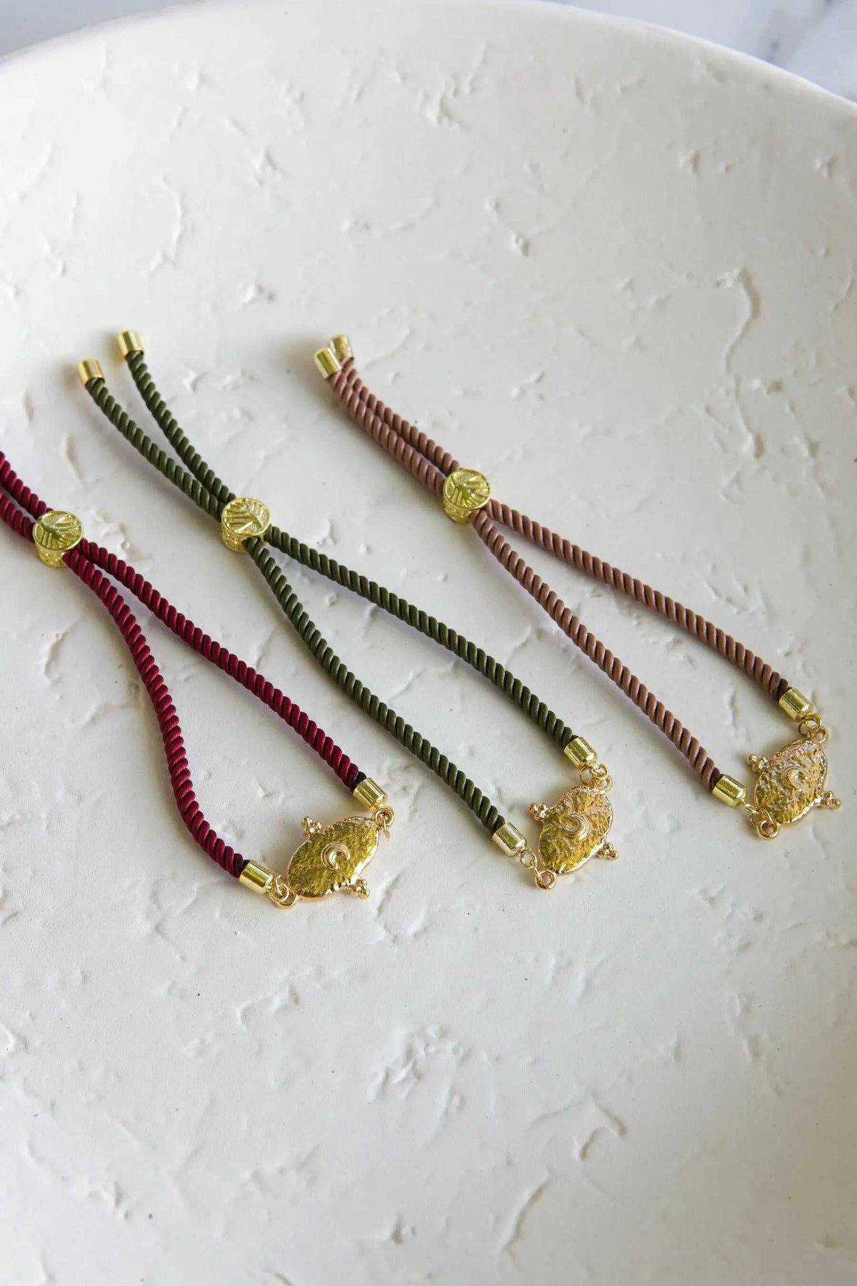 Adjustable rope Bracelet in burgundy, olive green, and dusty rose cords, each with a gold-tone medallion and charm