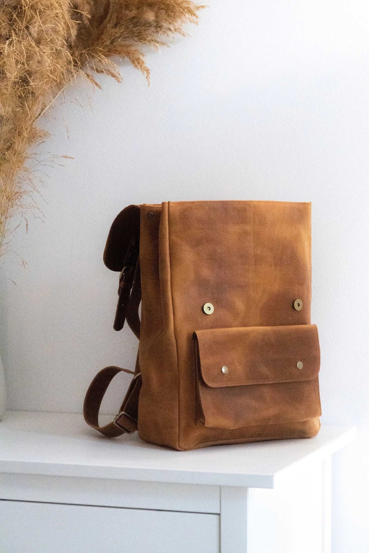 ALKION BACKPACK in mustard brown leather with a structured shape, large front flap pocket, and metal fasteners