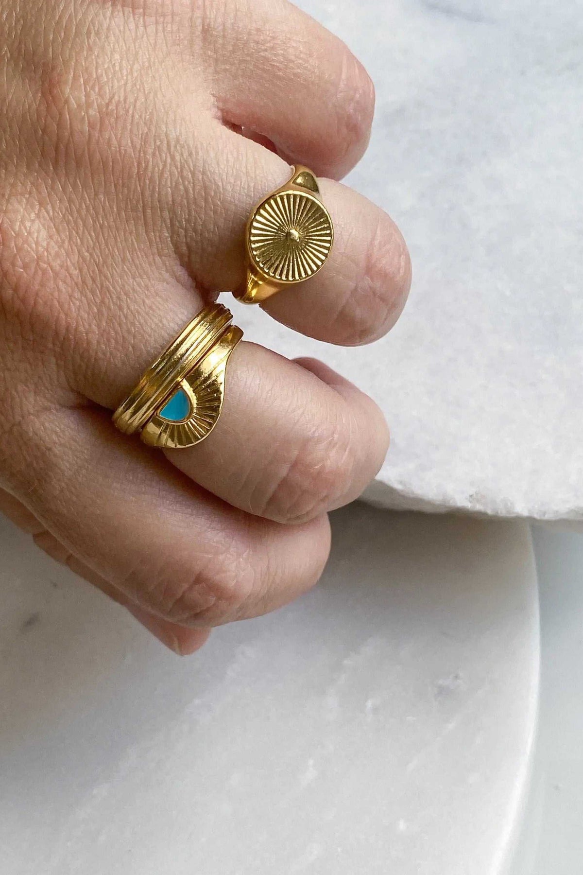 DELOS RING in gold-tone metal with a ridged circular sunburst face, shown beside a gold ring with a turquoise accent