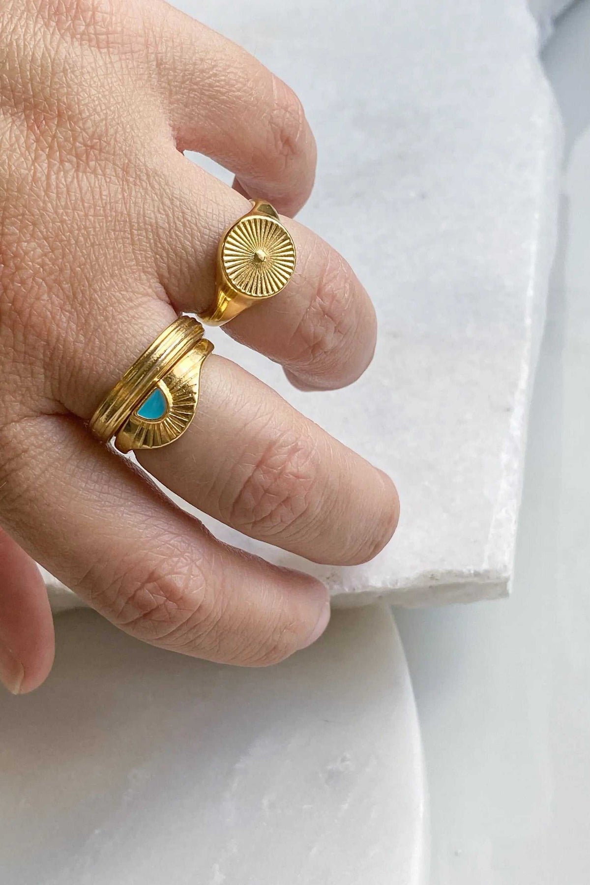 DELOS RING gold-tone sunburst signet ring with a round ridged face, shown beside a gold-tone ring with a turquoise drop