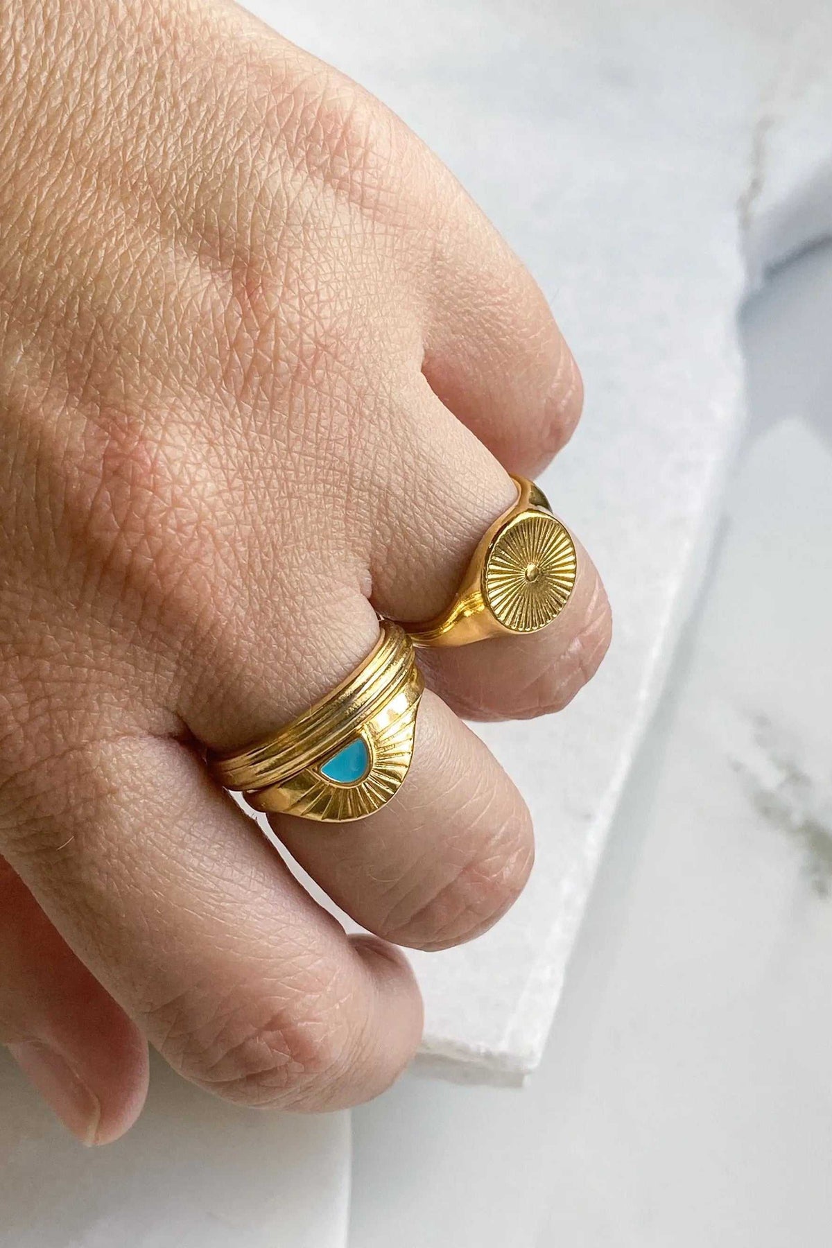 DELOS RING in gold-tone metal with a raised sunburst medallion, shown beside a second gold ring with a turquoise accent