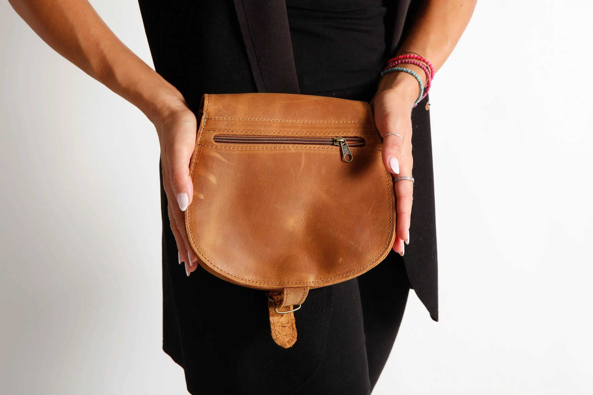 DIONE saddle shoulder bag in warm brown leather, with a rounded flap, front zipper pocket and metal buckle