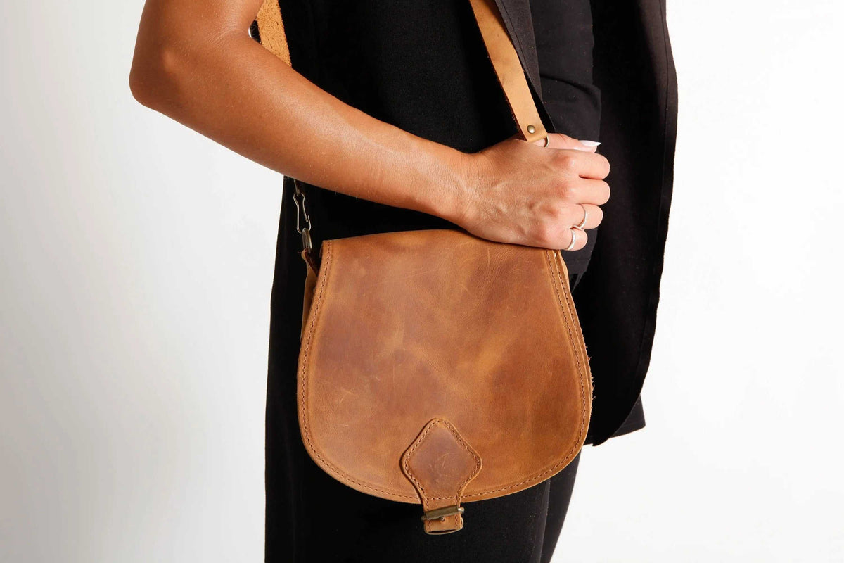 DIONE saddle shoulder bag in warm brown leather, featuring a rounded flap, central tab closure and slim strap