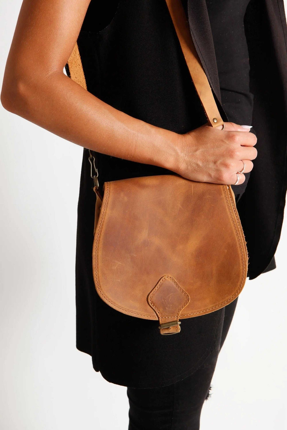 DIONE saddle shoulder bag in warm brown leather with a rounded flap, adjustable strap, and metal buckle closure