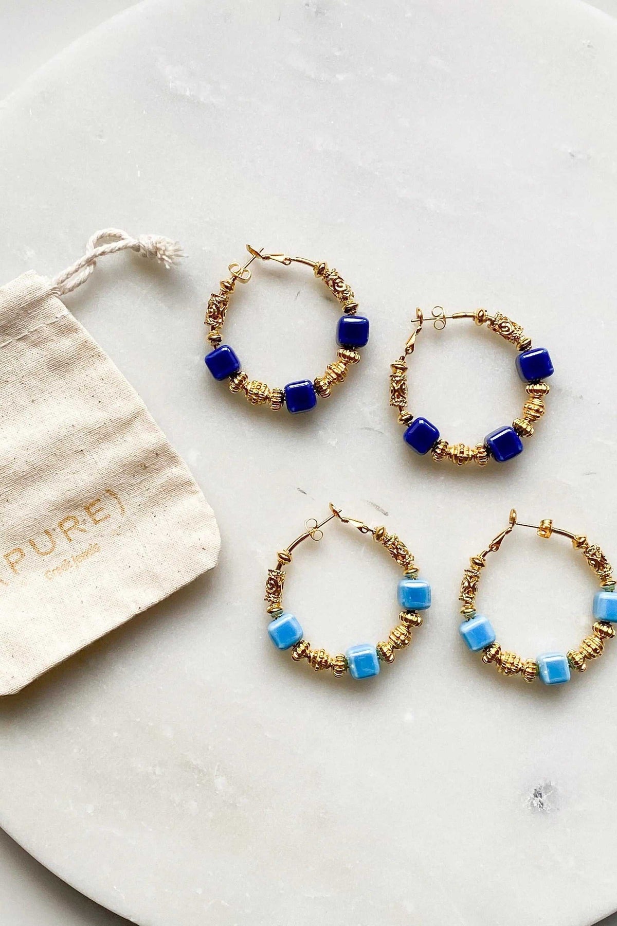 GÉA EARRINGS are gold-tone hoop earrings with cobalt blue or light blue ceramic cube beads and textured detailing