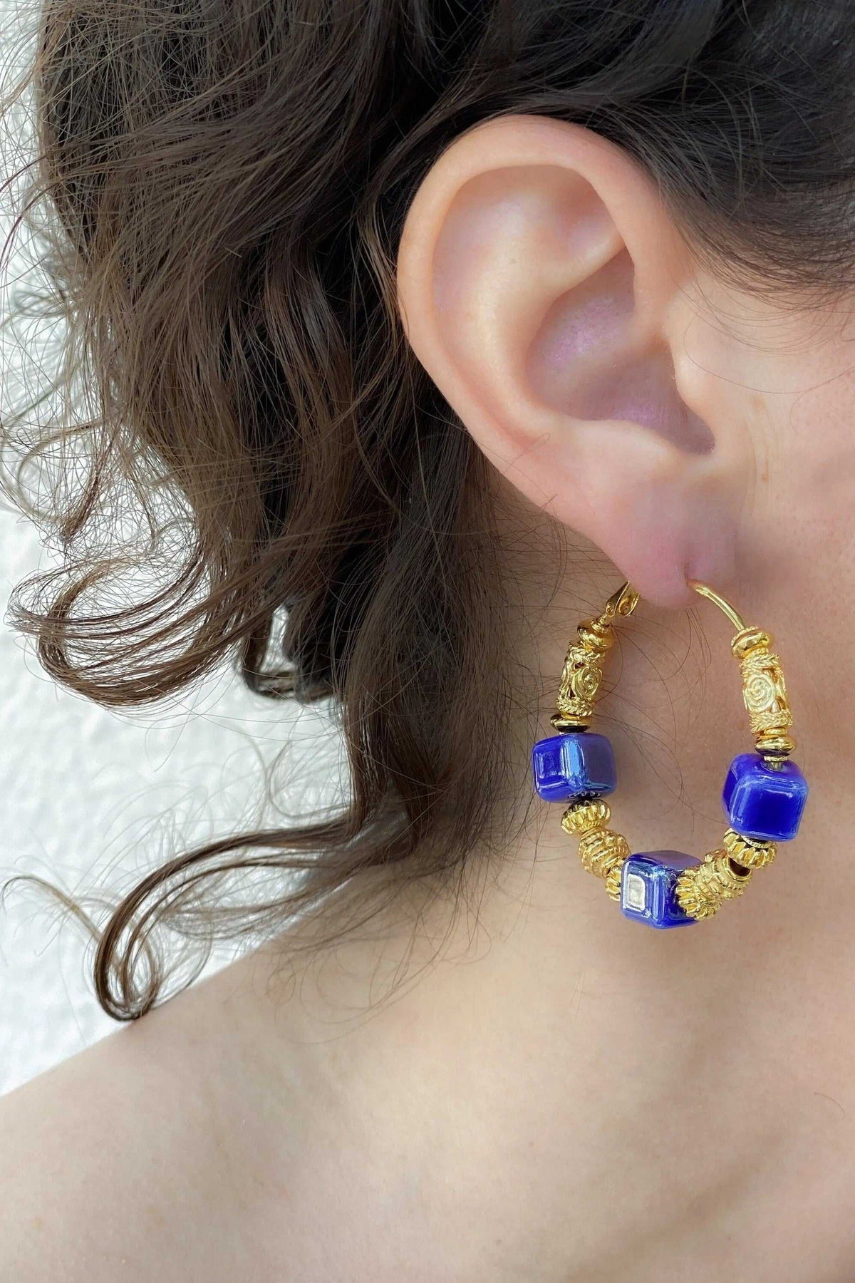 GÉA EARRINGS are gold-tone hoop earrings with vivid blue ceramic cube beads and ornate coiled metal details.