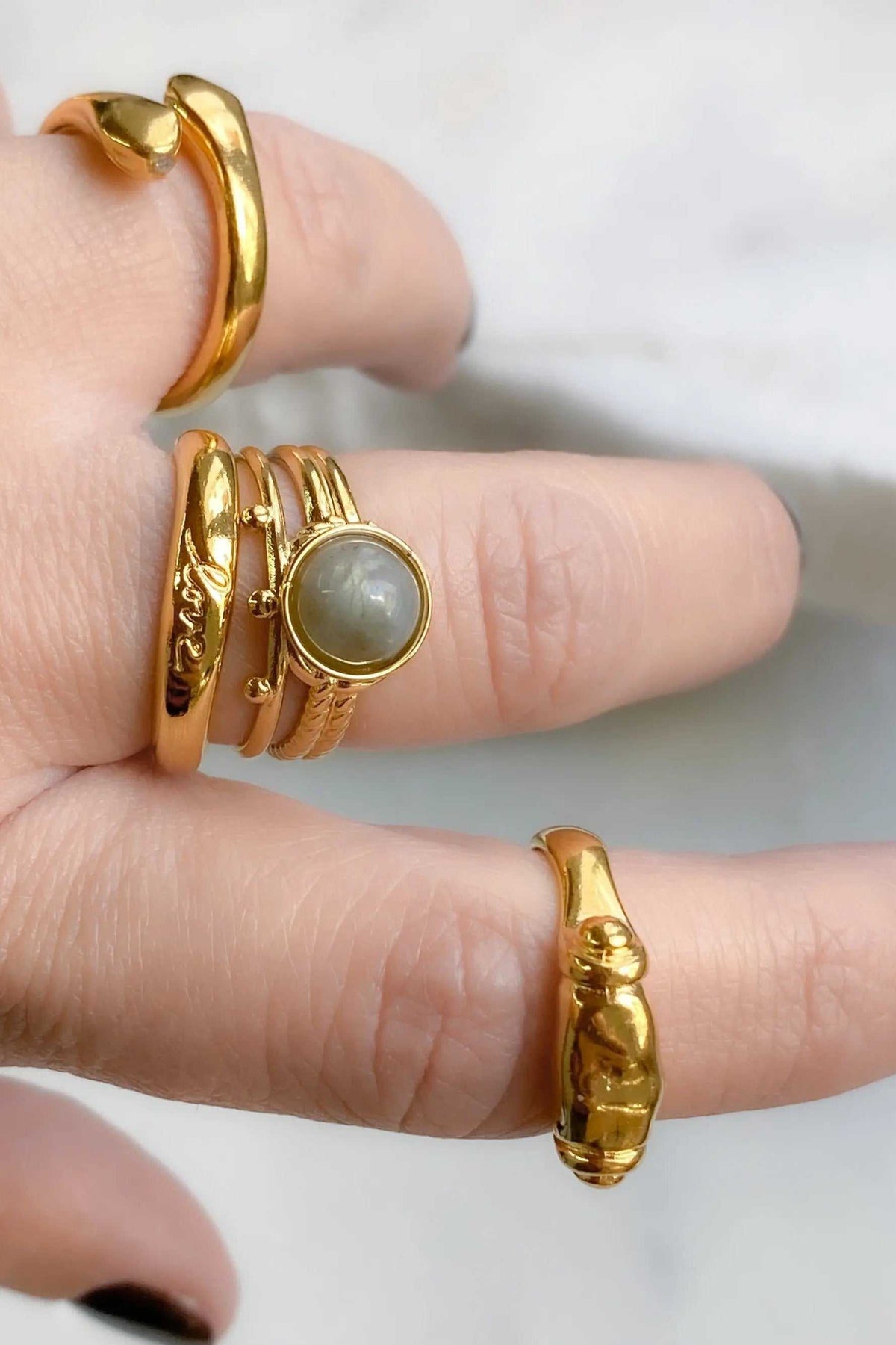 HERMINA Gold Stone Ring in gold-tone metal with a round grey-green stone, shown among sculpted, textured gold bands