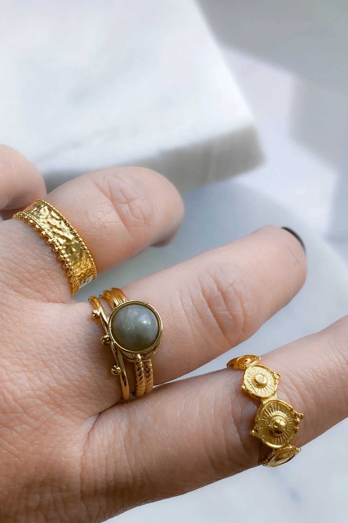 HERMINA Gold Stone Ring with a grey oval stone set in a textured gold-tone band, shown alongside ornate gold rings