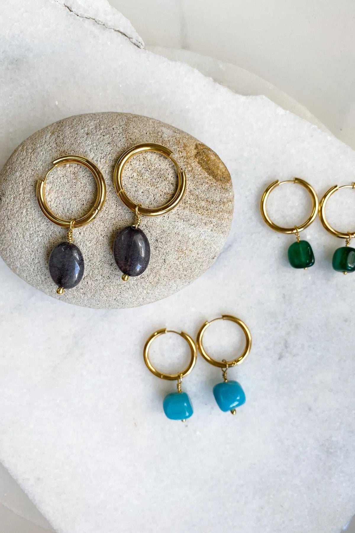 Large hoop earrings in gold-tone metal with dark blue oval stone charms, shown beside green and turquoise pairs
