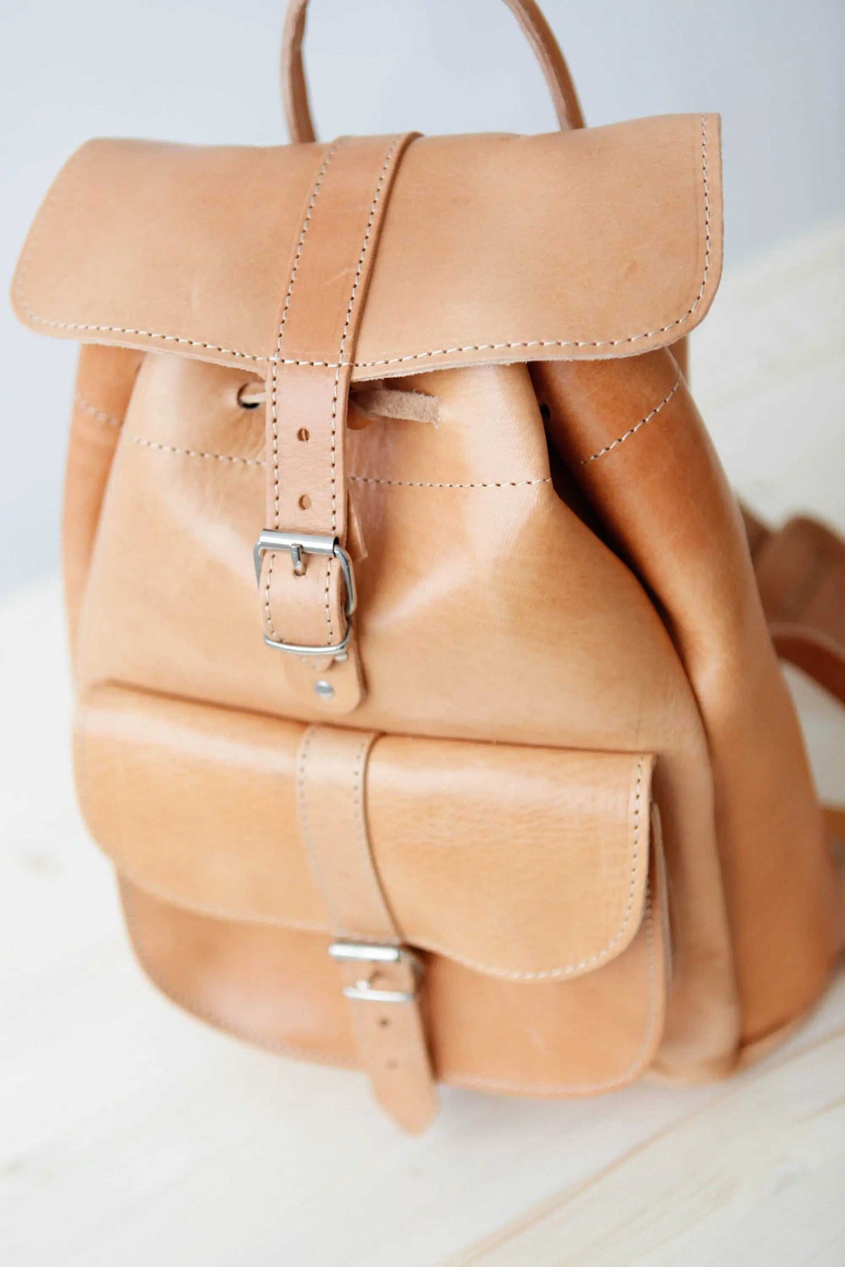 leather backpack in natural tan leather with stitched flap, drawstring closure, metal buckle, and large front pocket