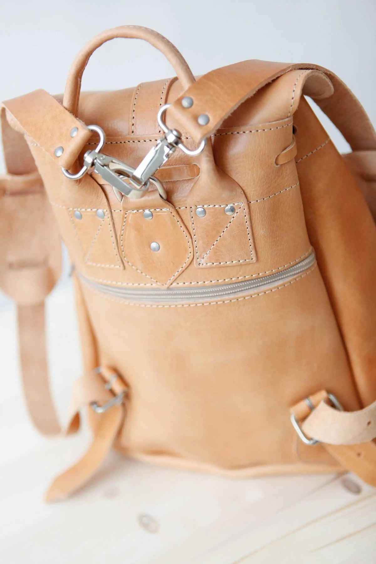 Leather backpack in natural tan leather with stitched front pockets, a metal buckle, drawstring closure and adjustable straps.