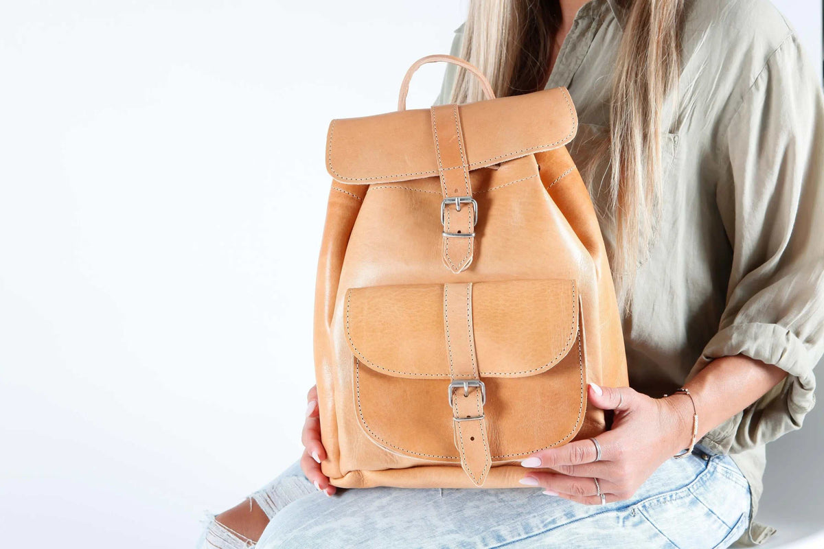 Leather backpack in warm tan leather with stitched flaps, two metal buckle closures, a top handle and front pocket