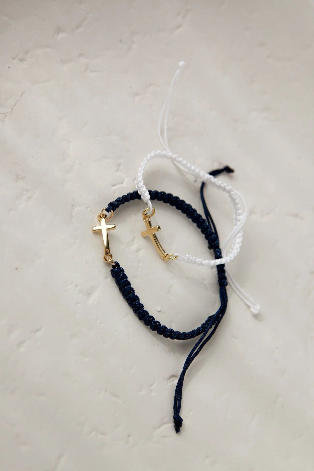 Macrame cross bracelet in dark blue and white woven cord, featuring gold-tone cross charms and adjustable sliding knots