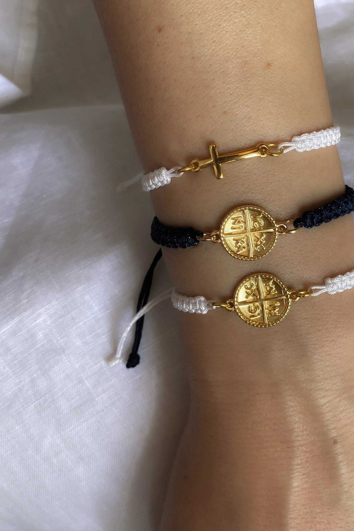 Macrame cross bracelet with white braided cord and a gold-tone cross charm, shown beside white and navy Byzantine coin bracelets.