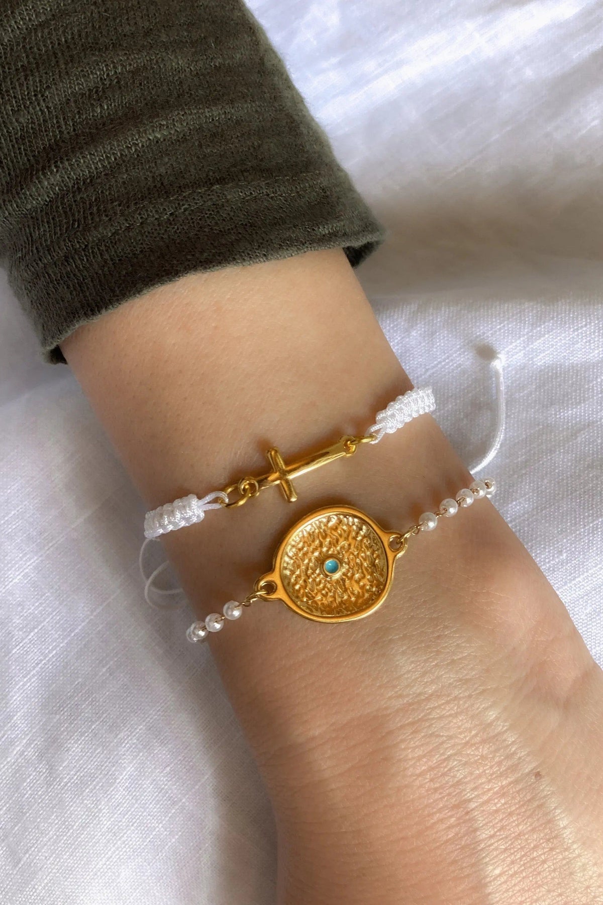 Macrame cross bracelet in white cord with gold-tone cross charm, paired with a pearl strand and ornate round medallion