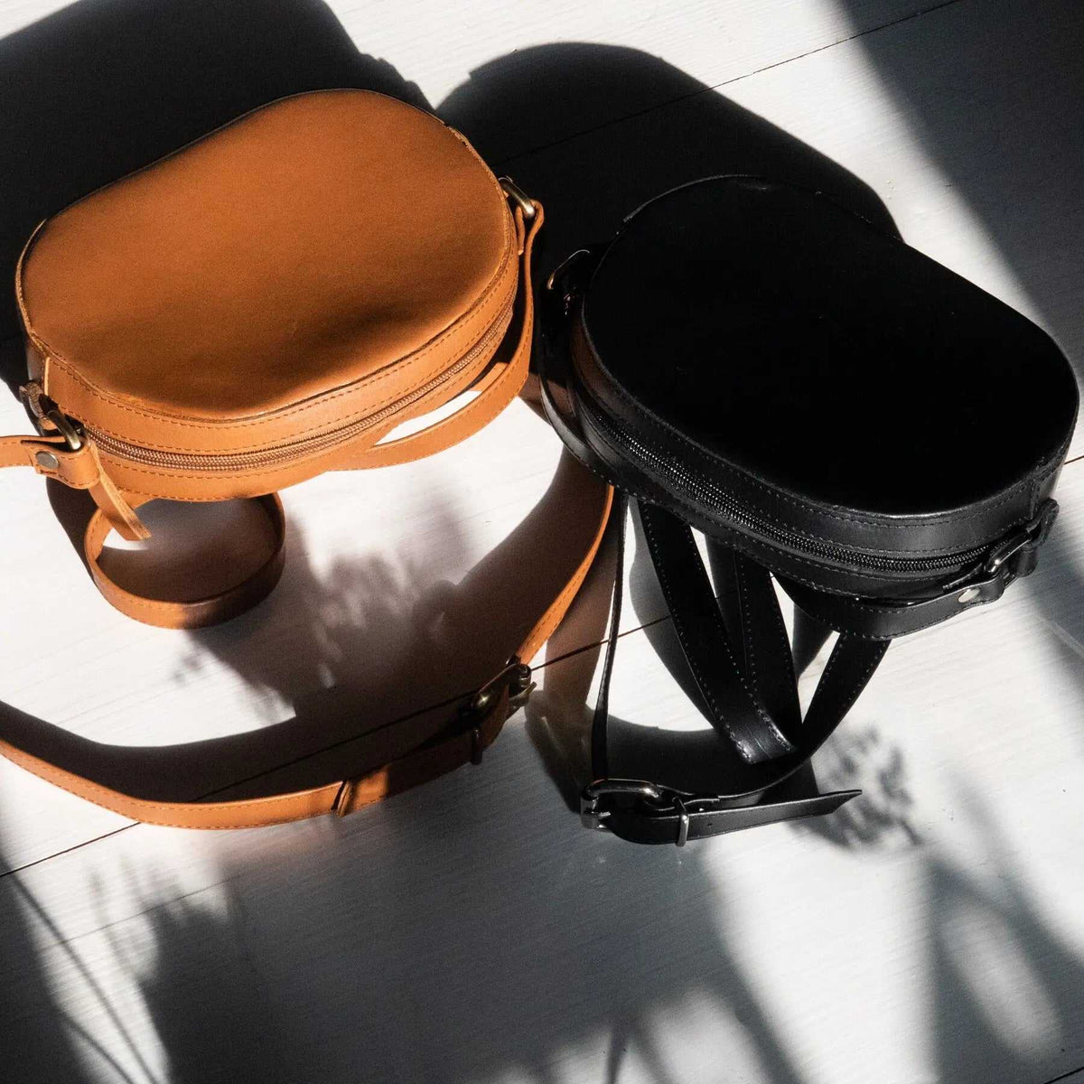 Minimal shoulder bag in tan and black leather with oval zip-top bodies, adjustable straps and metal buckles