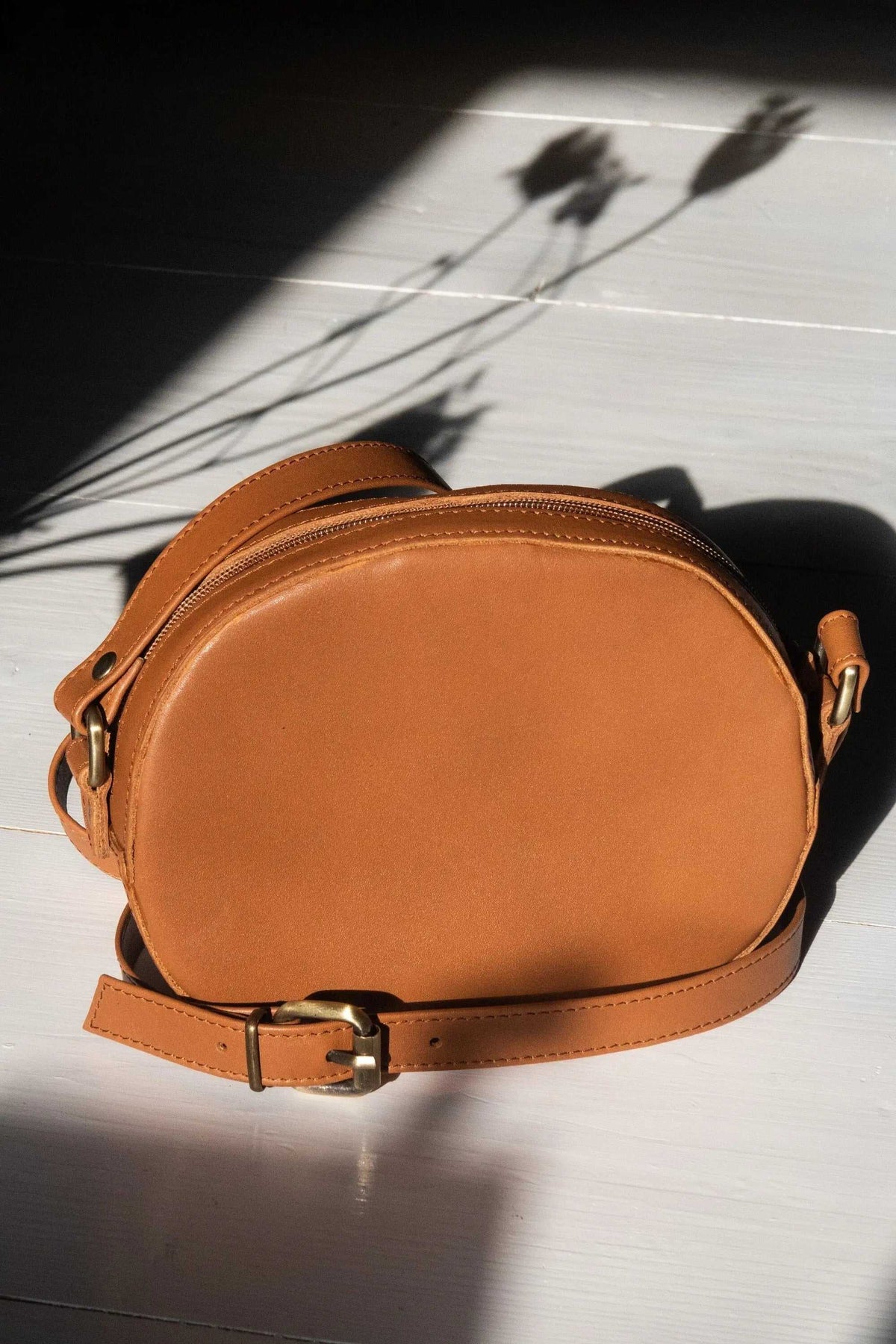minimal shoulder bag in warm tan leather with a smooth oval body, top zip, adjustable strap and gold-tone buckles