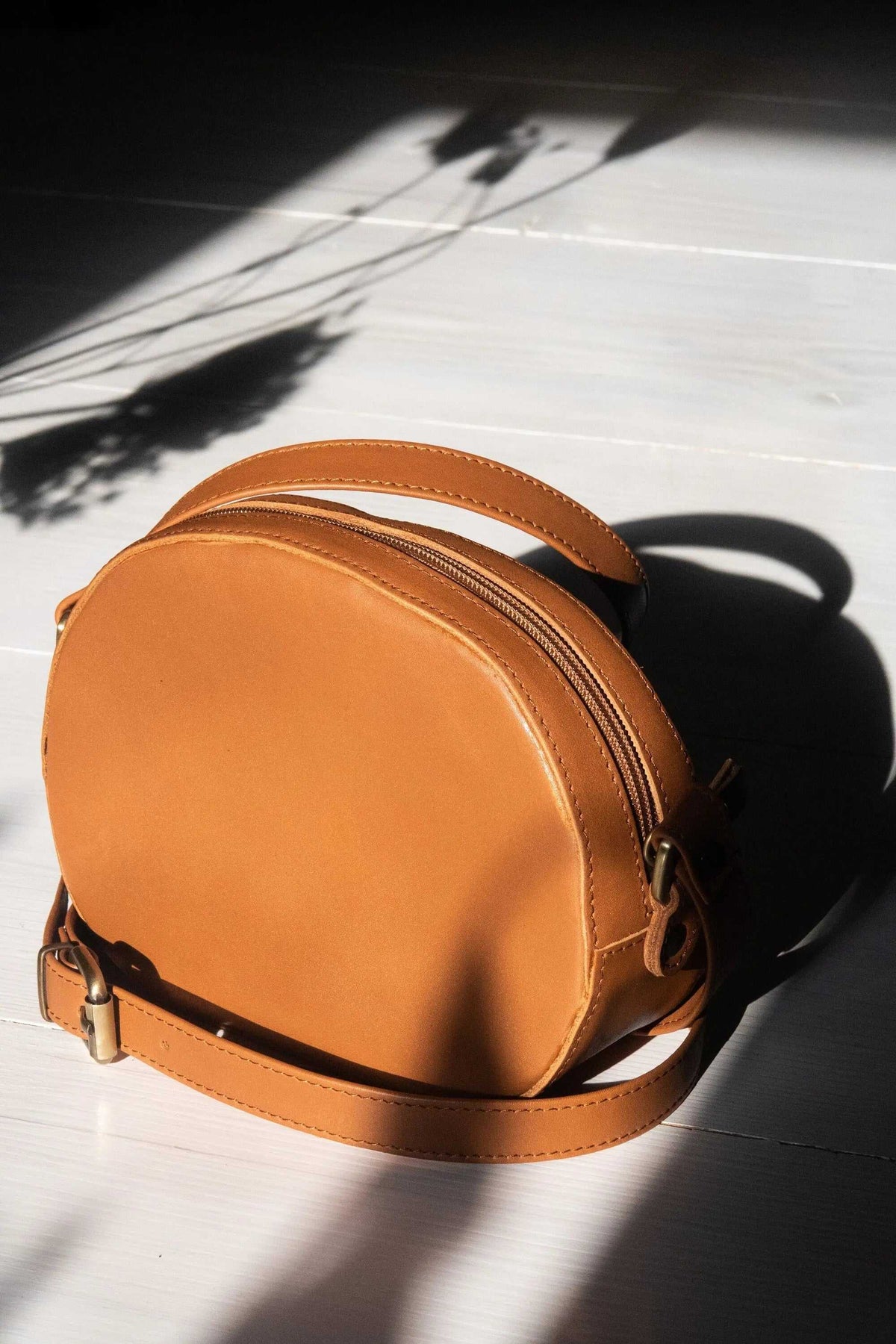 minimal shoulder bag in warm tan leather, with a rounded oval body, top zip, and adjustable strap with metal buckles
