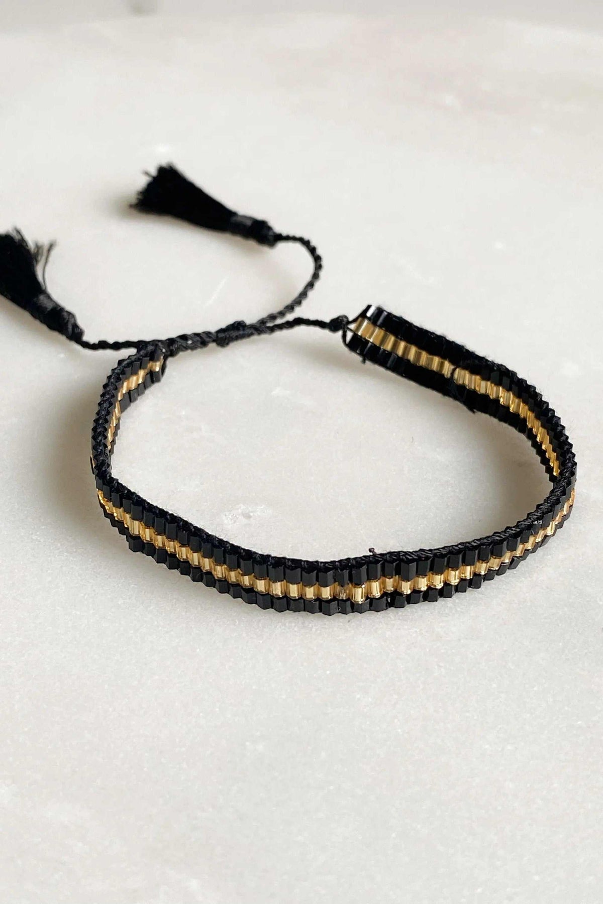 Miyuki beads bracelet woven with black thread, featuring a narrow gold bead stripe and adjustable tassel ties