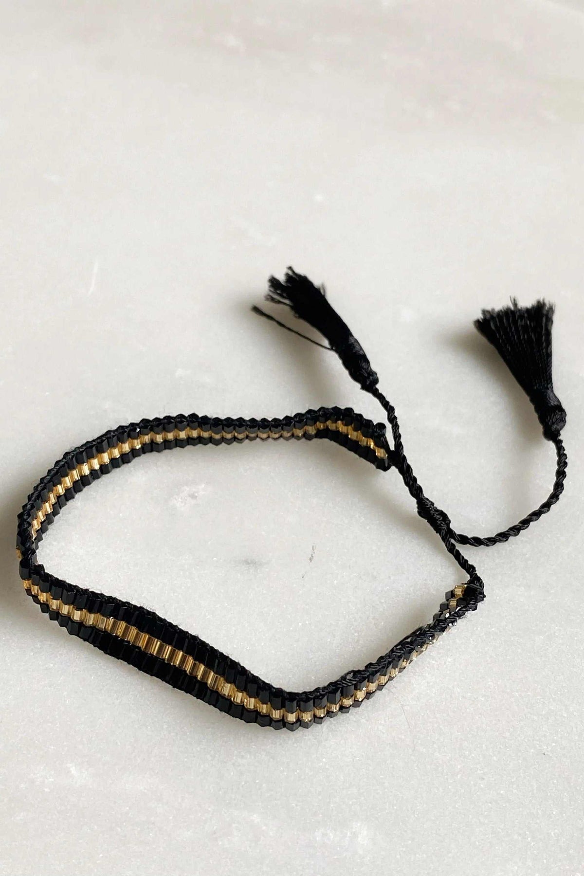 Miyuki beads bracelet woven in black fabric with a narrow gold glass bead stripe and adjustable tassel ties