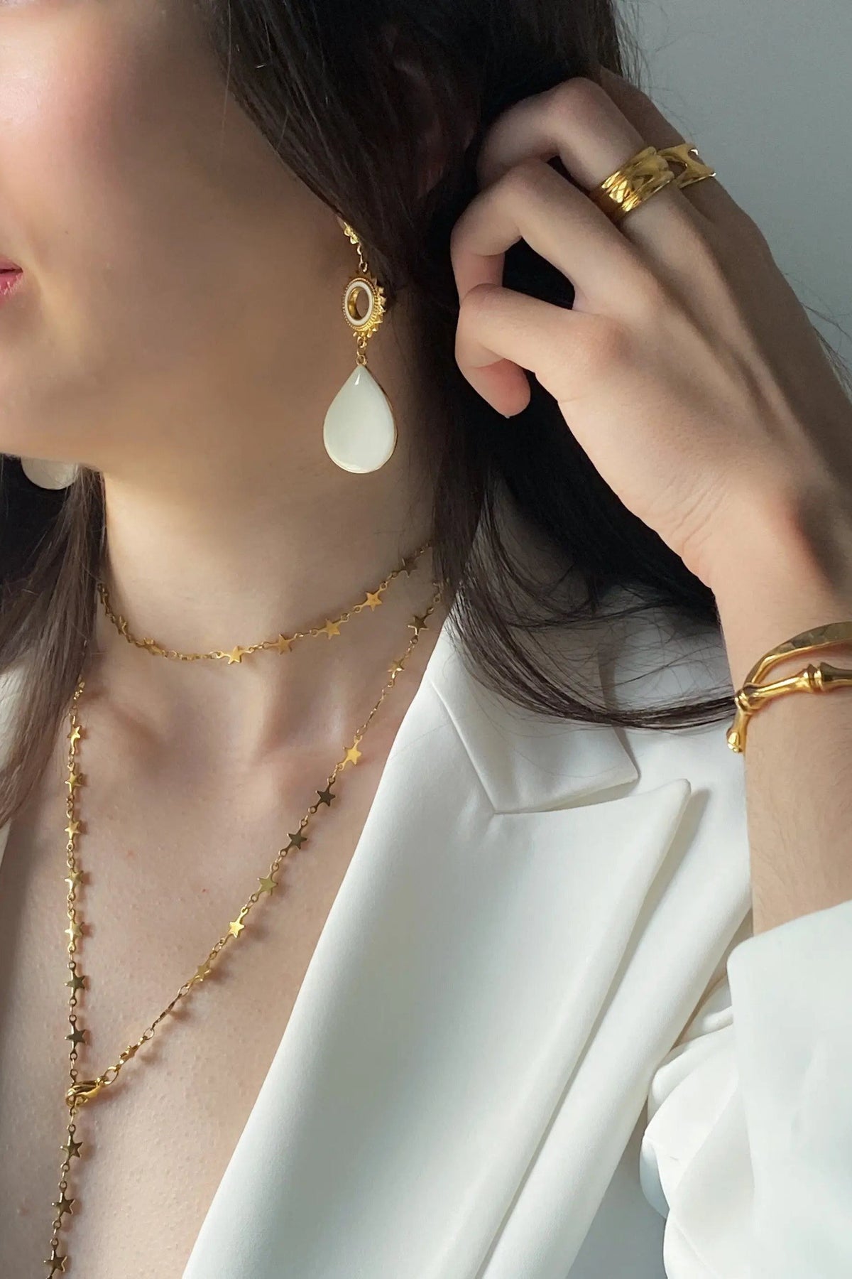 Sun drop earrings with gold-tone sunburst accents and smooth white teardrop pendants, worn with layered gold necklaces