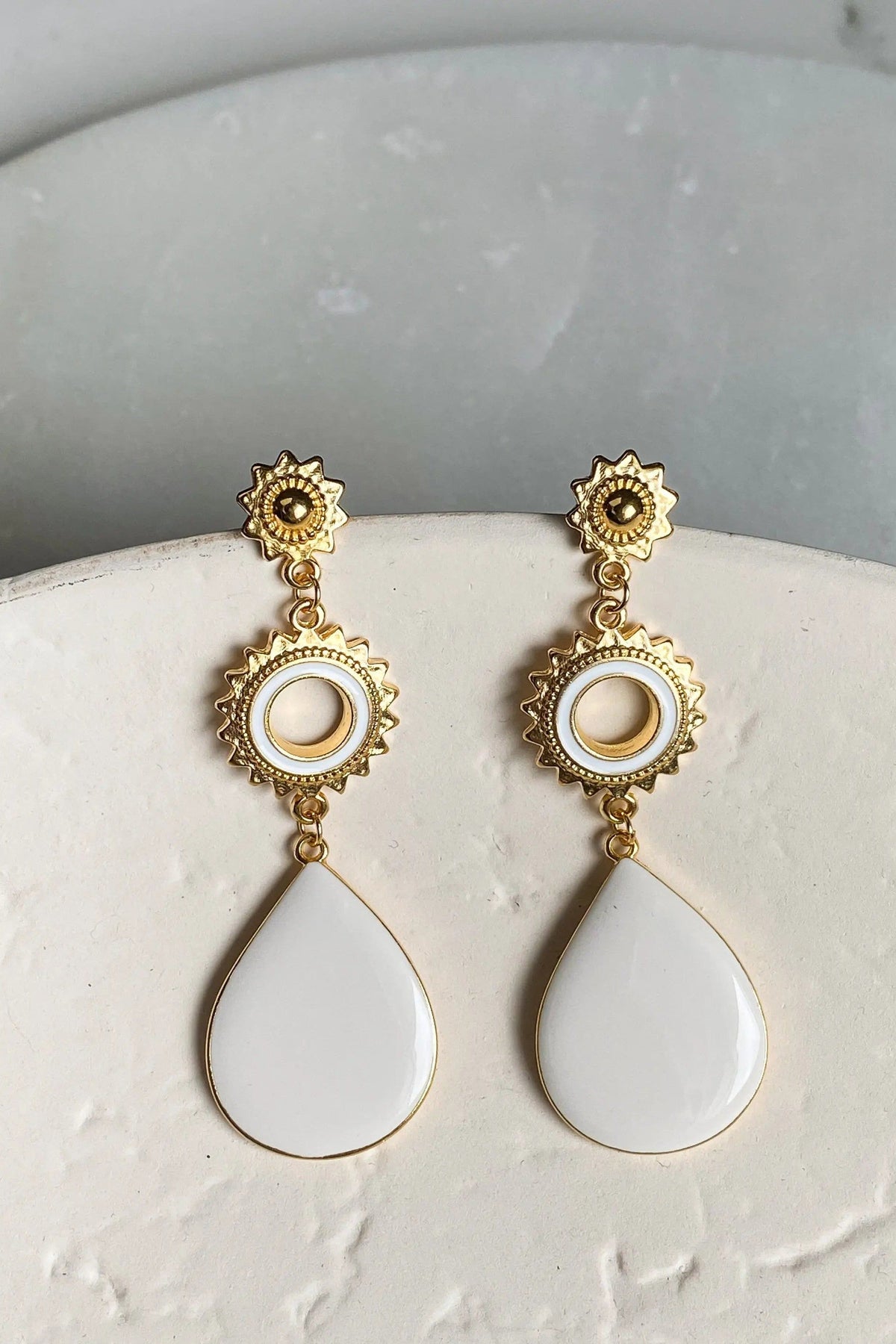 Sun drop earrings in gold-tone metal with glossy white teardrop pendants, circular ring details and sunburst accents