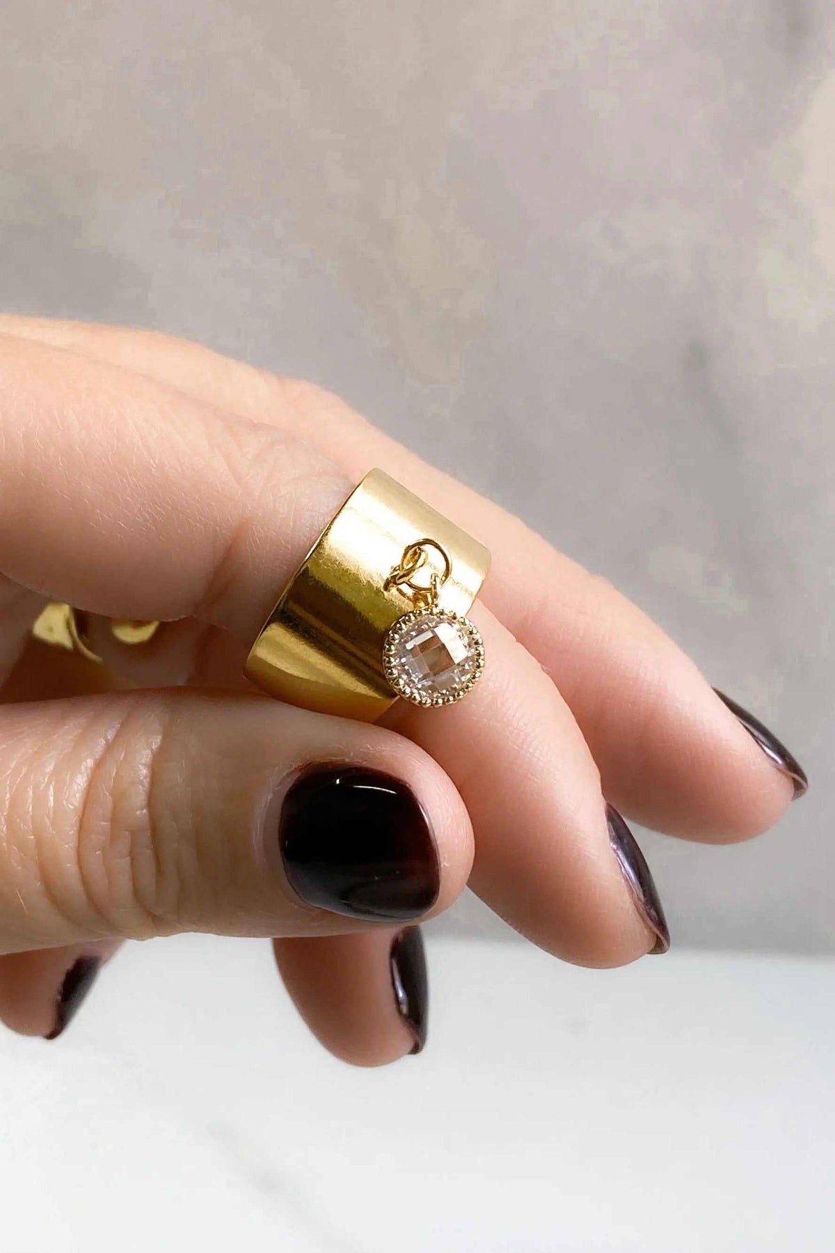 Wide flat band ring in polished gold with a dangling round clear stone charm and smooth tapered sides