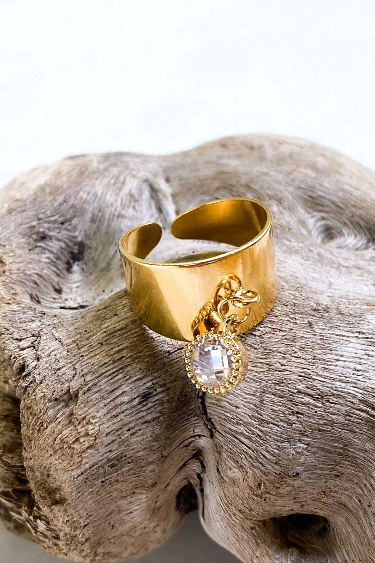 Wide flat band ring in gold-tone metal with a smooth adjustable band and dangling clear square-cut charm
