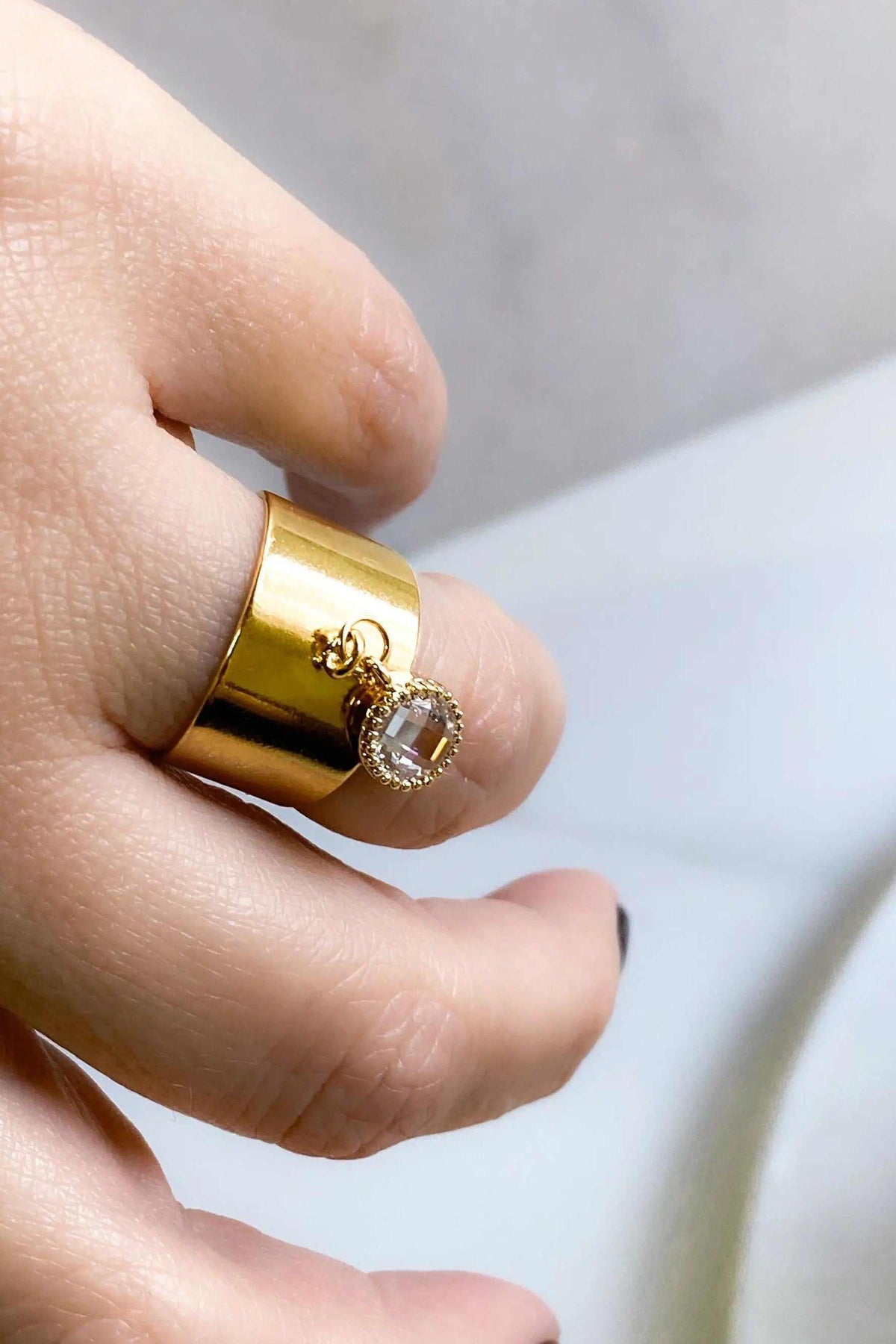 Wide flat band ring in polished gold-tone metal with a small clear oval stone charm and textured setting