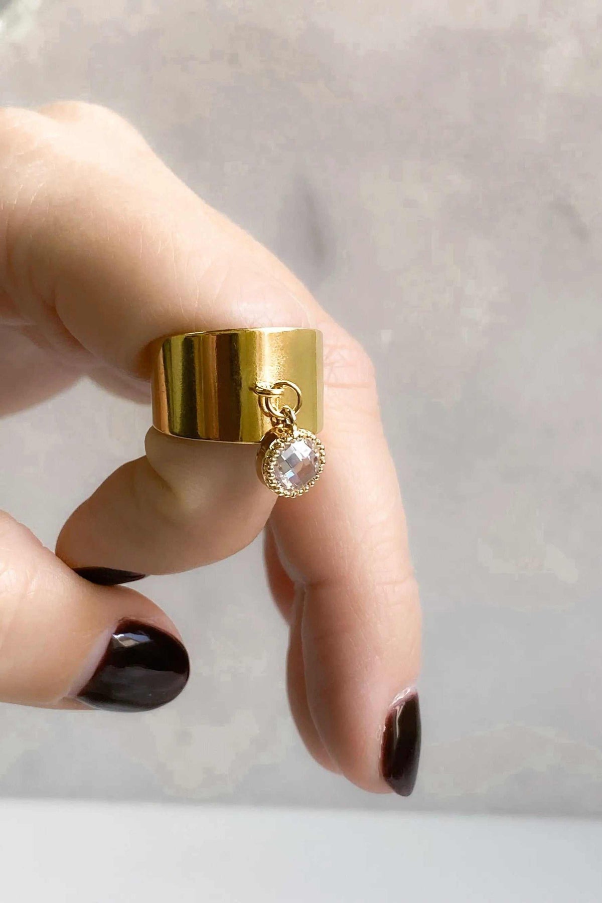 Wide flat band ring in polished gold-tone metal with a dangling round clear stone charm and a broad smooth band