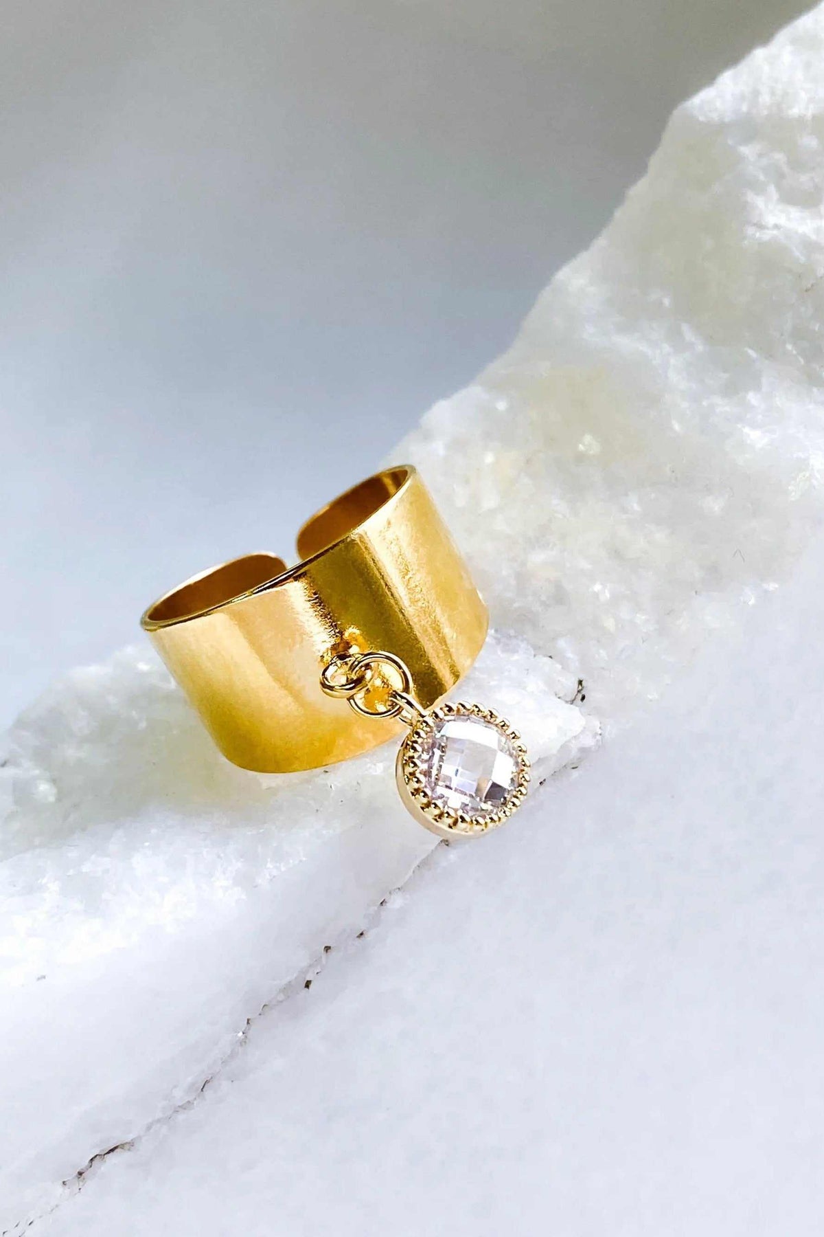Wide flat band ring in polished gold-tone metal with a broad smooth band and dangling clear square stone charm