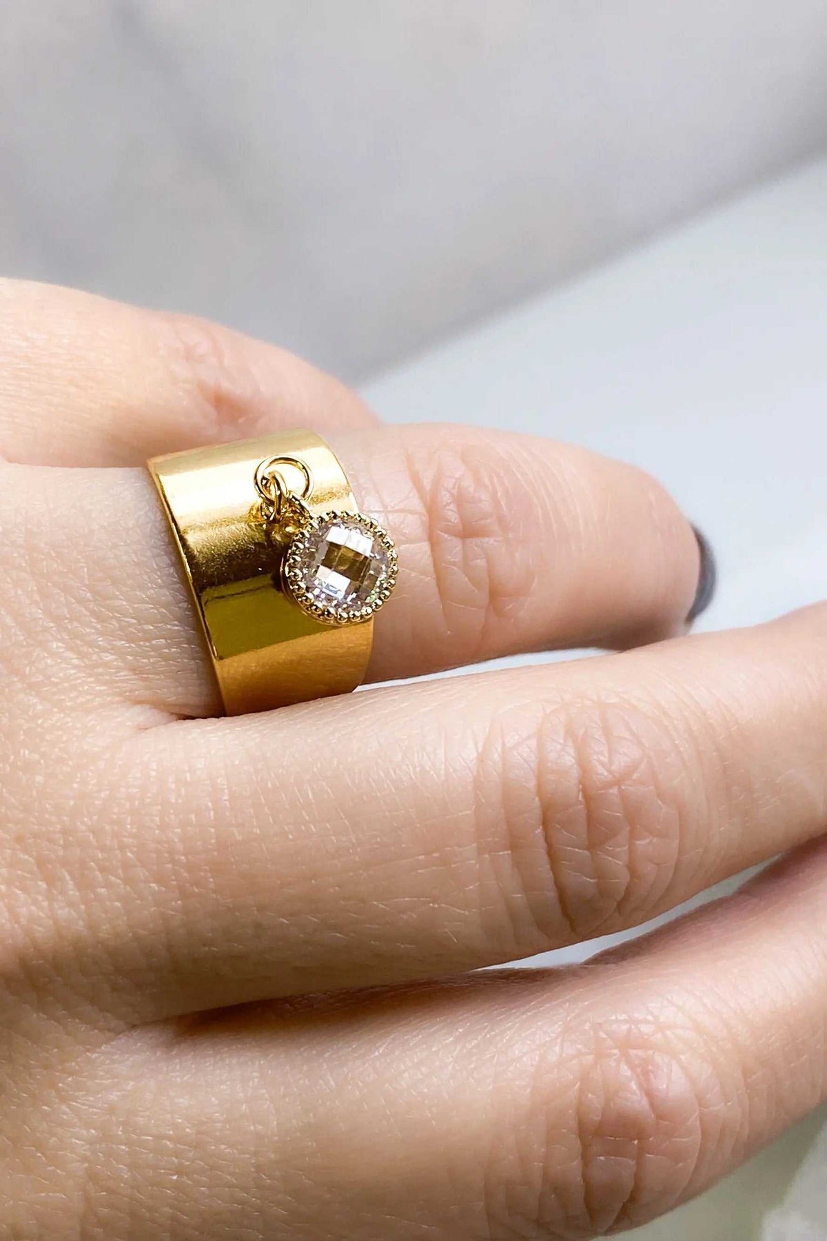 Wide flat band ring in polished gold-tone metal, featuring a round clear faceted charm suspended from a loop