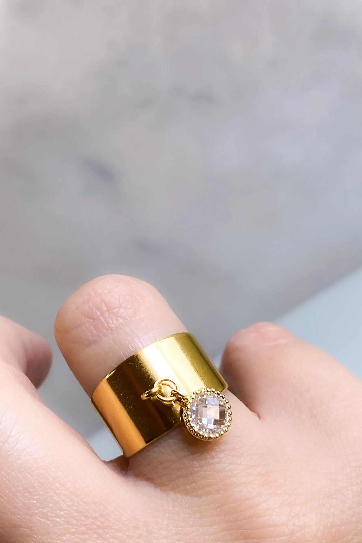 Wide flat band ring in polished gold-tone metal with a dangling clear square-cut stone charm framed by tiny accents
