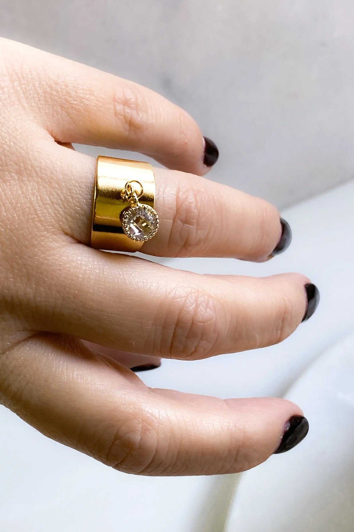 Wide flat band ring in polished gold tone, featuring a small round clear stone charm dangling from the band