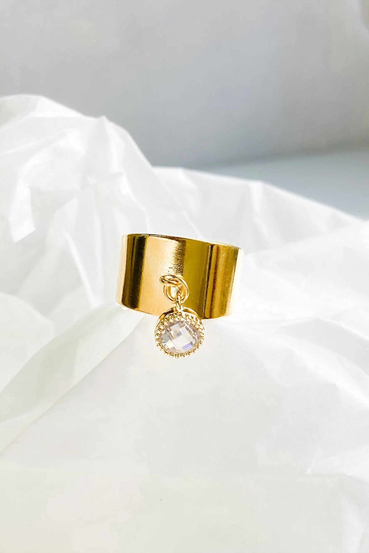 Wide flat band ring in polished gold-tone metal with a wide smooth band and a dangling round clear stone charm